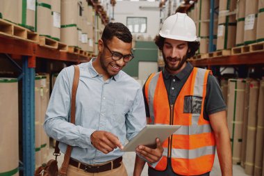 Young businessman showing data in touchpad to warehouse worker wearing hardhat and orange jacket uniform