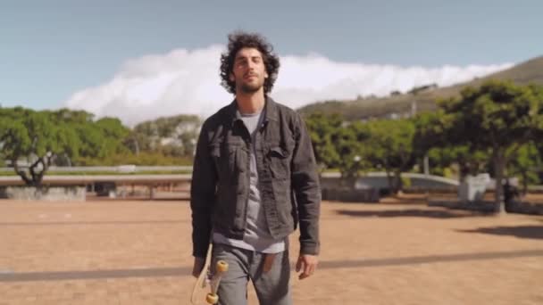 Retrato de un joven guapo deportivo feliz sosteniendo monopatín en la mano camina hacia la cámara sonriendo — Vídeo de stock