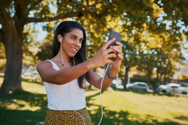 Sorrindo jovem na moda com fones de ouvido nos ouvidos tirando selfie no smartphone no parque sorrindo e feliz — Fotografia de Stock