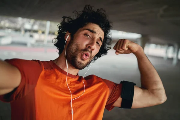 Attractive fit young sportsman with earphone making funny face flexing his muscles while taking a selfie outdoors — Stok fotoğraf