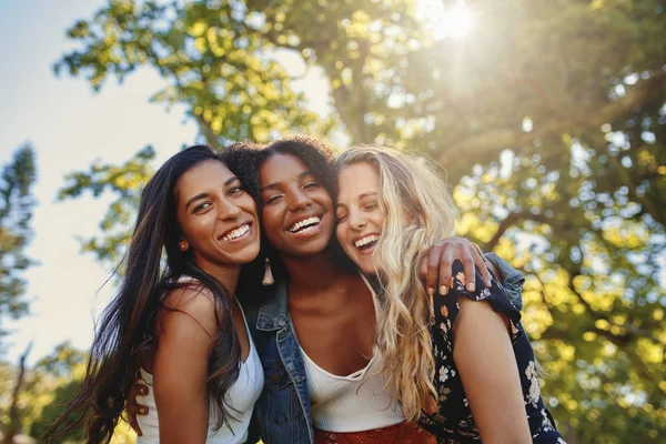 Tres mujeres jóvenes multiétnicas sonrientes en ropa casual de pie juntas bajo la brillante luz del sol en el parque divirtiéndose y riendo — Foto de Stock