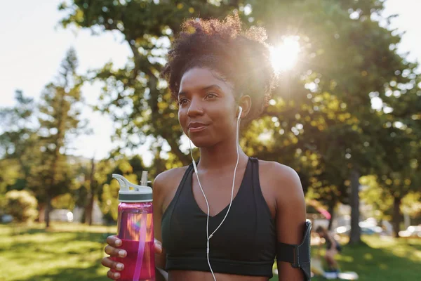 Mujer atlética parada en un día soleado brillante en el parque con auriculares en sus oídos sosteniendo una botella de agua potable ecológica en la mano en el parque — Foto de Stock