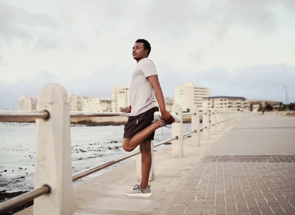 Vista lateral de un atleta joven que estira las piernas de pie junto a la barandilla en el paseo marítimo —  Fotos de Stock