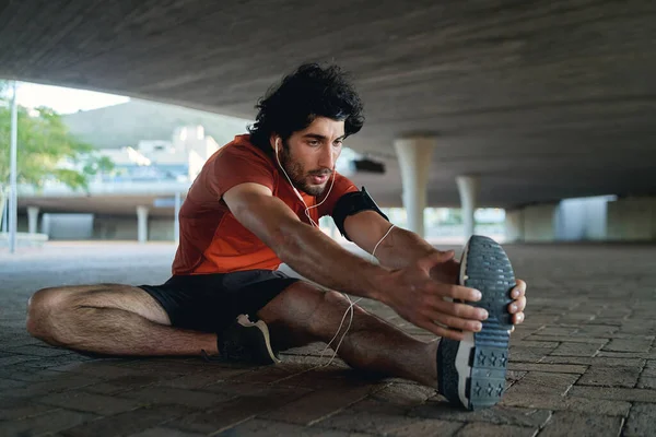 Junger männlicher Läufer hört Musik über Kopfhörer, streckt die Beine, bevor er läuft und auf der Straße unter der Brücke trainiert — Stockfoto