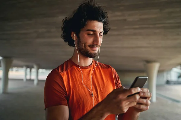 Портрет усміхненого молодого спортивного чоловіка, зайнятого в наборі на смартфон, що стоїть під мостом — стокове фото