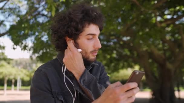 Portré egy modern, mosolygós fiatalemberről, aki fülhallgatót helyez a fülébe, miközben mobilon SMS-ezik a parkban. — Stock videók