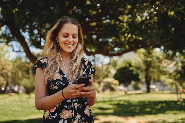 Портрет щасливої блондинки, яка насолоджується смс на мобільному телефоні в парку протягом сонячного дня, посміхаючись — стокове фото