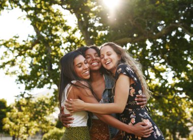 Portrait of happy three multiethnic multiracial female friends closely hugging and showing care and love for each other in park