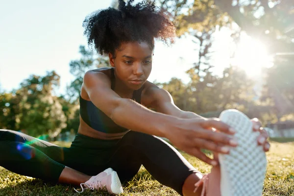 Portret van een Afrikaans-Amerikaanse jonge vrouw die in het park zit en haar benen uitstrekt in fel zonlicht - jonge zwarte vrouw die haar benen opwarmt voordat ze gaat sporten — Stockfoto