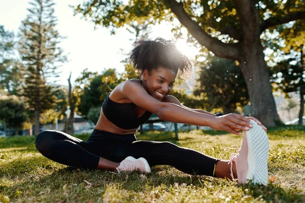 Glimlachend portret van een sportieve fit Afrikaans-Amerikaanse jonge vrouw zittend op gazon strekken haar benen in het park - gelukkig jonge zwarte vrouw opwarmen van haar spieren voor het lopen — Stockfoto