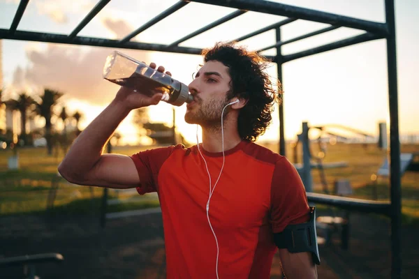 Deportivo joven caucásico hombre escuchando música en los auriculares agua potable de botella reutilizable transparente en el parque de gimnasio al aire libre — Foto de Stock