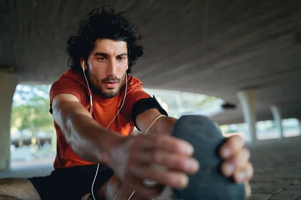 Retrato de un joven atleta deportivo saludable estirándose y calentándose antes de correr bajo el puente urbano - primer plano de un corredor masculino serio —  Fotos de Stock