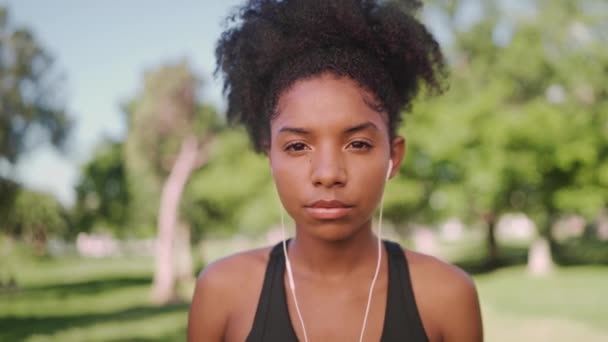 Πορτρέτο της ταιριάζει νεαρή αποφασισμένη Αφροαμερικανή γυναίκα ακούγοντας μουσική στα ακουστικά κοιτάζοντας στην κάμερα — Αρχείο Βίντεο