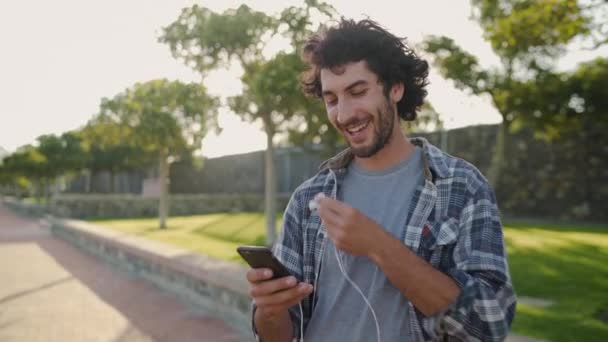 Усміхнений портрет молодого чоловіка, який вставляє навушники в вуха та смс онлайн на мобільний телефон у парку — стокове відео