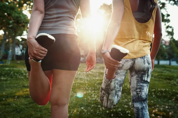 Primo piano di due giovani donne sportive che allungano le gambe nella giornata di sole nel parco - due donne multirazziali che allungano i quadricipiti prima di una corsa — Foto Stock
