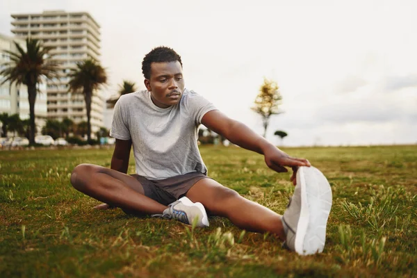 En afrikansk amerikansk ung fitness mand sidder på grønt græs strækker benene før træning - Stock-foto