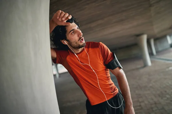 Lelah olahraga pemuda atletis dengan earphone dan armband bersandar pada kolom di bawah jembatan beristirahat setelah berlari atlet kelelahan beristirahat — Stok Foto