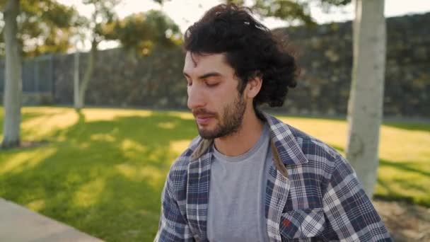 Крупним планом голодний красивий молодий чоловік сидить в парку їсть бутерброд під час спекотного літа — стокове відео