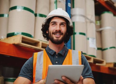 Envanter ve stokları kontrol etmek için elinde dijital tablet tutarken gülümseyen ve çalışan başarılı depo yöneticisi beyaz kask ve güvenlik ceketi giyiyor.