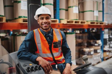 Koruyucu üniforma giyen yakışıklı mühendis ve kameraya sevinçle gülümsüyor forklift rafında çalışırken depoda oturuyor.