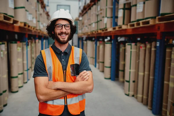 Cheerful male worker entrepreneur in uniform with white hardhat and arms crossed standing in from of warehouse rack arrangement wearing spectacles — Stock Photo, Image