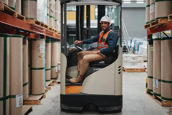 Smiling forklift driver wearing white helmet and vest sitting in machine transporting goods — Stockfoto