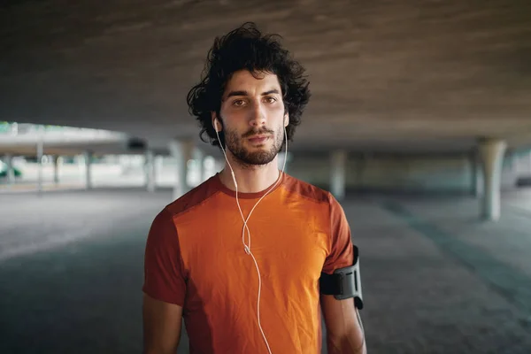 Retrato de jovem em forma grave com fones de ouvido em seu ouvido de pé sob a ponte olhando para a câmera — Fotografia de Stock