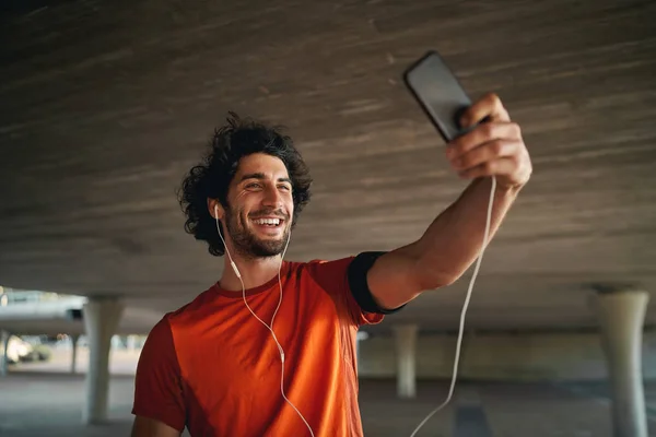 Jovem alegre de pé sob a ponte fazendo videochamada em seu smartphone com fone de ouvido - jovem homem apto a tirar uma selfie durante o exercício — Fotografia de Stock