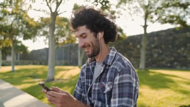 Вид збоку щасливий і спокійний красивий молодий чоловік насолоджується прослуховуванням музики на навушниках через мобільний телефон у парку — стокове відео