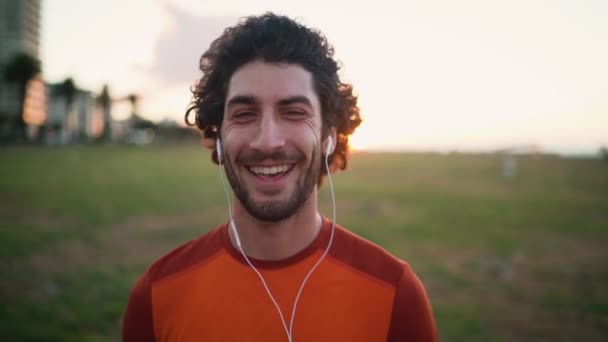 Primer plano del corredor masculino en auriculares mirando seriamente a la cámara durante el entrenamiento matutino — Vídeos de Stock