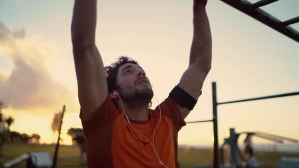 Jeune athlète sportif homme avec des écouteurs dans ses oreilles faire des pull-ups sur la barre horizontale à l'aire de jeux — Video