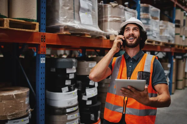 Knappe gelukkige man in witte helm en veiligheidsvest staan met digitale tablet terwijl praten via de telefoon weg te kijken tussen goederen in gangpad — Stockfoto