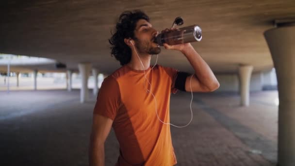 Sonriente hombre de fitness cansado bebe agua de una botella transparente después de trotar en la ciudad — Vídeo de stock