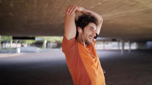 Портрет улыбающегося спортсмена, слушающего музыку в наушниках, делающего упражнения на растяжку и разогревающегося под мостом — стоковое видео