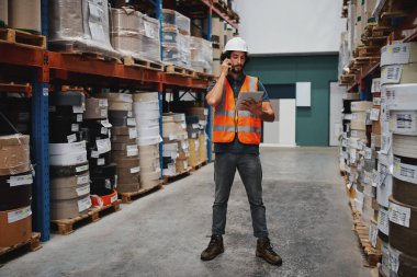 Full length of warehouse manager tracking supply order details using digital tablet while in conversation over mobile phone in warehouse standing between cardboard boxes in shelf