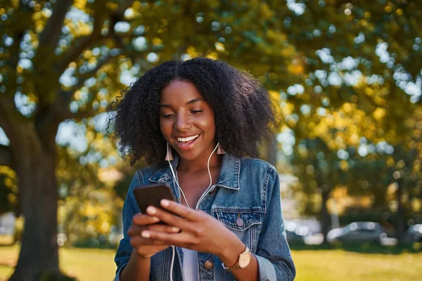 Portrait of smiling young african american woman listening to music on earphone using mobile phone in the park - Happy young black woman listening to music in the park on a sunny day — Stock Photo, Image