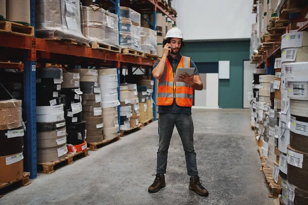 Full length of warehouse manager tracking supply order details using digital tablet while in conversation over mobile phone in warehouse standing between cardboard boxes in shelf — Stockfoto