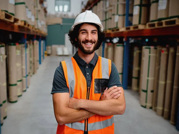 Portrait of cheerful supervisor in a warehouse for delivering and transporting industrial goods wearing white helmet and orange vest uniform standing with folded arms in aisle — Stockfoto
