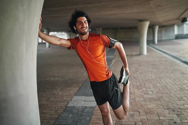 Retrato de um jovem sorridente que gosta de música em seu fone de ouvido aquecendo antes de correr sob a ponte — Fotografia de Stock