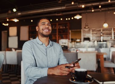 Handsome young african man laughing while reading funny message on phone sitting in modern cafeteria during free time