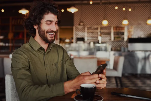 Jovem bonito alegre relaxante no café moderno usando celular enquanto bebe café — Fotografia de Stock