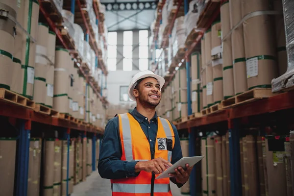 Low angle view of young african man wearing reflective jacket holding digital tablet standing in factory warehouse smiling — Stockfoto