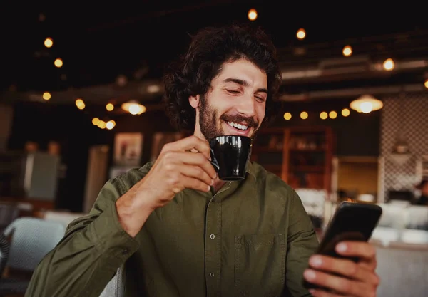 Handsome cheerful caucasian man using mobile phone while drinking coffee in modern cafeteria — Stockfoto