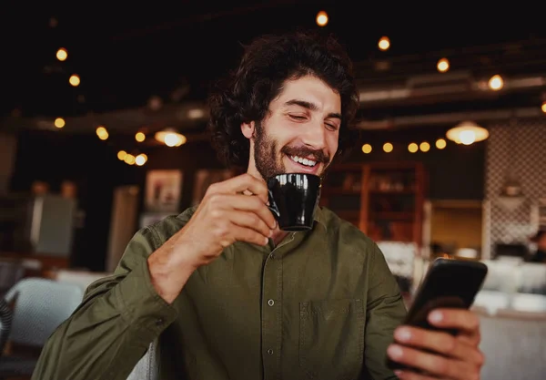 Bonito homem caucasiano alegre usando telefone celular enquanto bebe café na cafetaria moderna — Fotografia de Stock