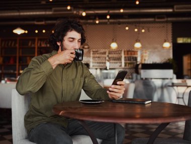 Handsome young caucasian man browsing through smartphone while drinking coffee in cafeteria