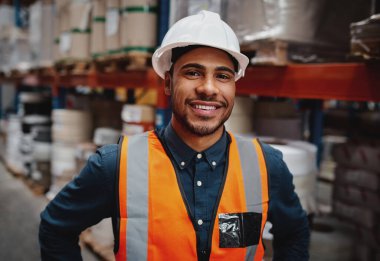 Happy warehouse manager standing in factory wearing white helmet and orange safety vest looking at camera smiling