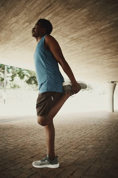 Hombre de fitness en ropa deportiva haciendo ejercicio al aire libre estirando su pierna mientras está de pie debajo del puente —  Fotos de Stock