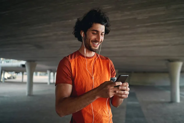 Portrait of a smiling young male runner standing under the concrete bridge with earphone in his ears typing on a smart phone