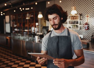 Genç kafe sahibi, kafede siyah önlükle dikilirken dijital tablet siparişini kontrol ediyor.