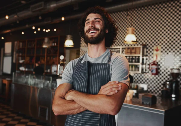 Vrolijke jongeman draagt schort staande met gevouwen armen in koffiehuis — Stockfoto